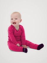 A smiling baby sitting on a light gray studio background, wearing a bright pink long-sleeve footed sleeper and dark purple socks. #color_peak pink