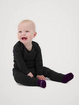 A baby sitting on a light gray studio background, wearing a black long-sleeve top and black pants with bright purple socks. #color_teton eclipse