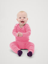 A baby sitting and smiling, wearing a bright pink long-sleeve footed sleeper with dark purple socks, photographed against a clean light gray background. #color_alpenglow