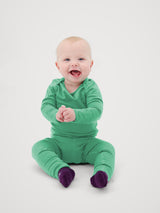 A baby sitting and smiling, wearing a bright green long-sleeve footed one-piece pajama with dark purple socks, photographed against a seamless light gray background. #color_meadow