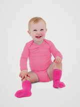A smiling baby sitting on a light gray studio background, wearing a bright pink long-sleeve bodysuit and matching pink socks. #color_alpenglow