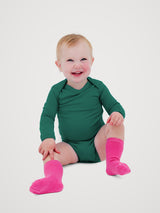 A smiling baby sitting on a light gray studio background, wearing a long-sleeve dark green bodysuit and bright pink socks. #color_evergreen