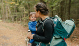 A woman hiking on a forest trail carrying a toddler in a teal and gray baby carrier backpack. The woman wears a black long-sleeve top, and the baby is dressed in a blue outfit.