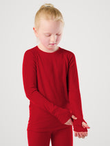 A young girl with blonde hair in a ponytail wearing a bright red long-sleeve shirt and matching red leggings, standing against a light gray studio background. #color_campfire