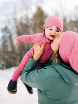 A baby in a pink one-piece outfit and matching hat is being held up outdoors by an adult wearing a green jacket and a pink hood. The background shows a snowy outdoor setting with blurred trees. #color_alpenglow