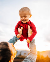 Dad swinging a laughing toddler upside down outdoors at sunset, toddler wearing a red Merino Wool Adventure Zippy romper #color_campfire