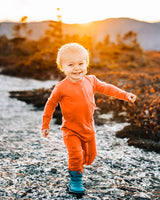 Toddler running along a rocky creek shoreline at sunset, wearing an orange-red Merino Wool Adventure Zippy romper and boots #color_poppy