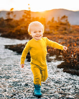 Toddler running along a rocky creek shoreline at sunset, wearing a golden yellow Merino Wool Adventure Zippy romper and boots #color_yellowstone