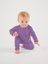A baby with light hair wearing a solid purple long-sleeve zip-up sleeper, kneeling on a white studio background. #color_lilac