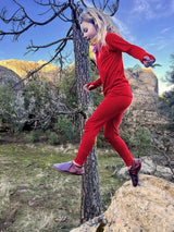 A young girl wearing a bright red Iksplor merino wool long-sleeve base layer top and matching leggings jumping on rocks. #color_campfire