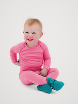 A smiling baby sitting cross-legged on a light gray studio background, wearing a bright pink long-sleeve one-piece outfit with teal booties. #color_alpenglow