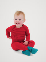 A smiling baby sitting cross-legged on a light gray studio background, wearing a bright red long-sleeve footless one-piece pajama or romper with teal socks. #color_campfire