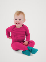A smiling baby sitting cross-legged on a light gray studio background, wearing a bright pink long-sleeve footless one-piece and teal booties. #color_peak pink
