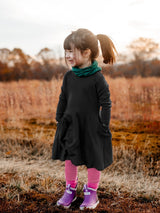 Young girl wearing an Iksplor black merino wool adventure dress with pockets, pink leggings, and purple rain boots. #color_teton eclipse - black