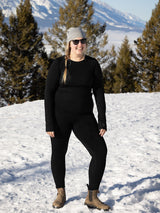 Woman wearing Iksplor black merino wool midweight leggings and matching long sleeve top in a snowy mountain setting. #color_teton eclipse