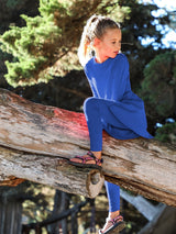 A young girl wearing a royal blue Iksplor merino wool adventure dress with leggings while climbing a fallen tree. #color_ocean