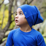 Child wearing a royal blue Iksplor merino wool beanie with a small mountain logo, outdoors in a forest setting. #color_ocean