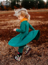 A young girl wearing an Iksplor green merino wool adventure dress with pockets, spinning in a field. #color_evergreen