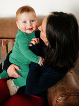 Iksplor baby wearing a green merino wool long sleeve shirt with fold-over mitts held by a mother on a leather sofa. #color_meadow