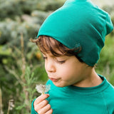 Child wearing a green Iksplor merino wool beanie while blowing on a dandelion in an outdoor setting. #color_evergreen