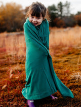 A young girl wrapped in a soft green Iksplor merino wool adventure blanket standing in a grassy field. #color_evergreen