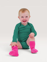 A smiling baby wearing an Iksplor long-sleeve green merino wool bodysuit and bright pink socks. #color_evergreen