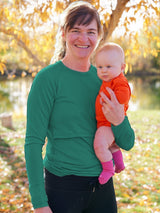 A mother wearing a green Iksplor merino wool long sleeve nursing top with thumb holes while holding her baby outdoors. #color_evergreen