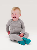Iksplor baby wearing grey merino wool long sleeve top and pants with teal booties, sitting and smiling. #color_trail dust