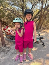 Two children wearing Iksplor magenta merino wool tank tops and matching shorts while biking in a sunny forest. #color_peak pink