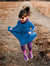 Iksplor kids merino wool adventure dress in blue worn by a girl jumping in a field with purple leggings and boots #color_ocean