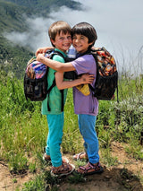 Two happy children wearing Iksplor merino wool joggers and t-shirts while hiking outdoors with backpacks. #color_meadow