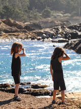 Two children wearing black Iksplor merino wool tank tops and shorts standing on a rocky coast by the ocean #color_teton eclipse