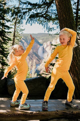 Two girls wearing bright yellow Iksplor merino wool long sleeve tops and leggings dancing outdoors near a waterfall.  #color_yellowstone