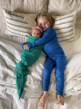 Iksplor merino wool adventure blanket used as a green swaddle for a baby next to a child in blue merino wool pajamas.