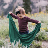 Iksplor Merino Wool Adventure Blanket in green held by a toddler in a mountain meadow #color_evergreen