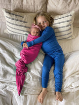 Iksplor Merino Wool Adventure Blanket used to swaddle a baby in pink, next to a child in blue merino wool pajamas. #color_peak pink