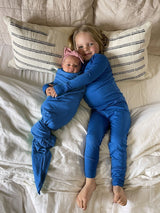 Iksplor Merino wool adventure blanket used to swaddle a baby next to a child in matching blue wool pajamas on a bed. #color_ocean