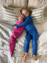 Iksplor Merino Wool Adventure Blanket used to swaddle a baby in pink, next to a child in blue merino wool pajamas. #color_peak pink