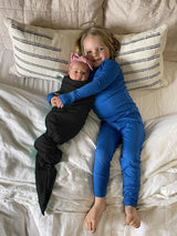 Iksplor merino wool adventure blanket used to swaddle a newborn baby next to a child in blue pajamas on a bed.