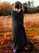 Iksplor Merino Wool Adventure Blanket wrapped around a smiling child standing in a grassy outdoor field #color_teton eclipse