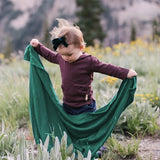 Iksplor Merino Wool Adventure Blanket in green held by a toddler in a mountain meadow #color_evergreen