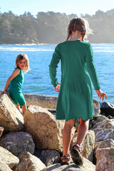 Iksplor kids merino wool adventure dress in forest green worn by a girl climbing rocks by the ocean #color_evergreen