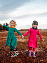 Two girls wearing Iksplor merino wool adventure dresses in green and pink while holding hands in a field. #color_evergreen