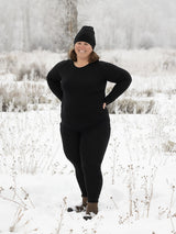 A woman wearing black Iksplor merino wool midweight leggings and matching long sleeve top in a snowy field. #color_teton eclipse
