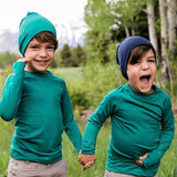 Two boys wearing Iksplor merino wool beanies in teal and navy blue while holding hands in a grassy field. #color_evergreen