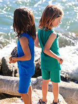 Two children wearing Iksplor merino wool adventure tank tops and shorts in blue and green by the ocean. #color_ocean