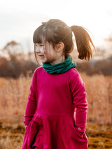 Young girl wearing a green Iksplor merino wool neck gaiter and a magenta long-sleeve dress outdoors. #color_evergreen
