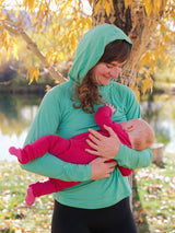 Iksplor merino wool nursing sun hoodie in green worn by a mother breastfeeding her baby outdoors in autumn #color_meadow