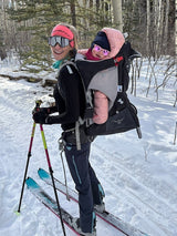 Iksplor merino wool nursing top worn by a mother cross-country skiing with a baby in a carrier on a snowy trail. #color_teton eclipse - black