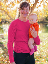 Iksplor merino wool nursing top in bright pink with thumb holes, worn by a mother holding her baby outdoors. #color_peak pink
